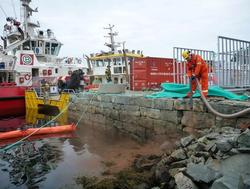 Mann i arbeidsklær holder slange ved bryggekanten nær to båter og en container. Overskyet vær. Fotografi.
