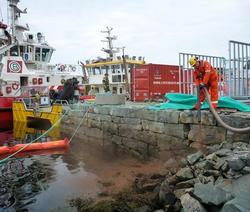 Mann i arbeidsklær holder slange ved bryggekanten nær to båter og en container. Overskyet vær. Fotografi.