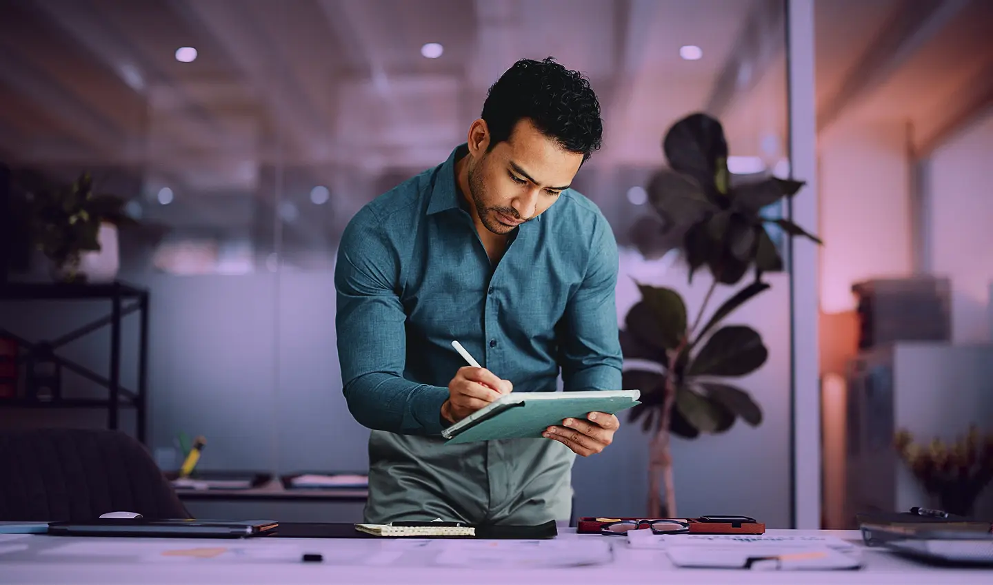 Homme écrivant sur une tablette dans un bureau moderne, concentré sur son travail devant une table remplie de documents.
