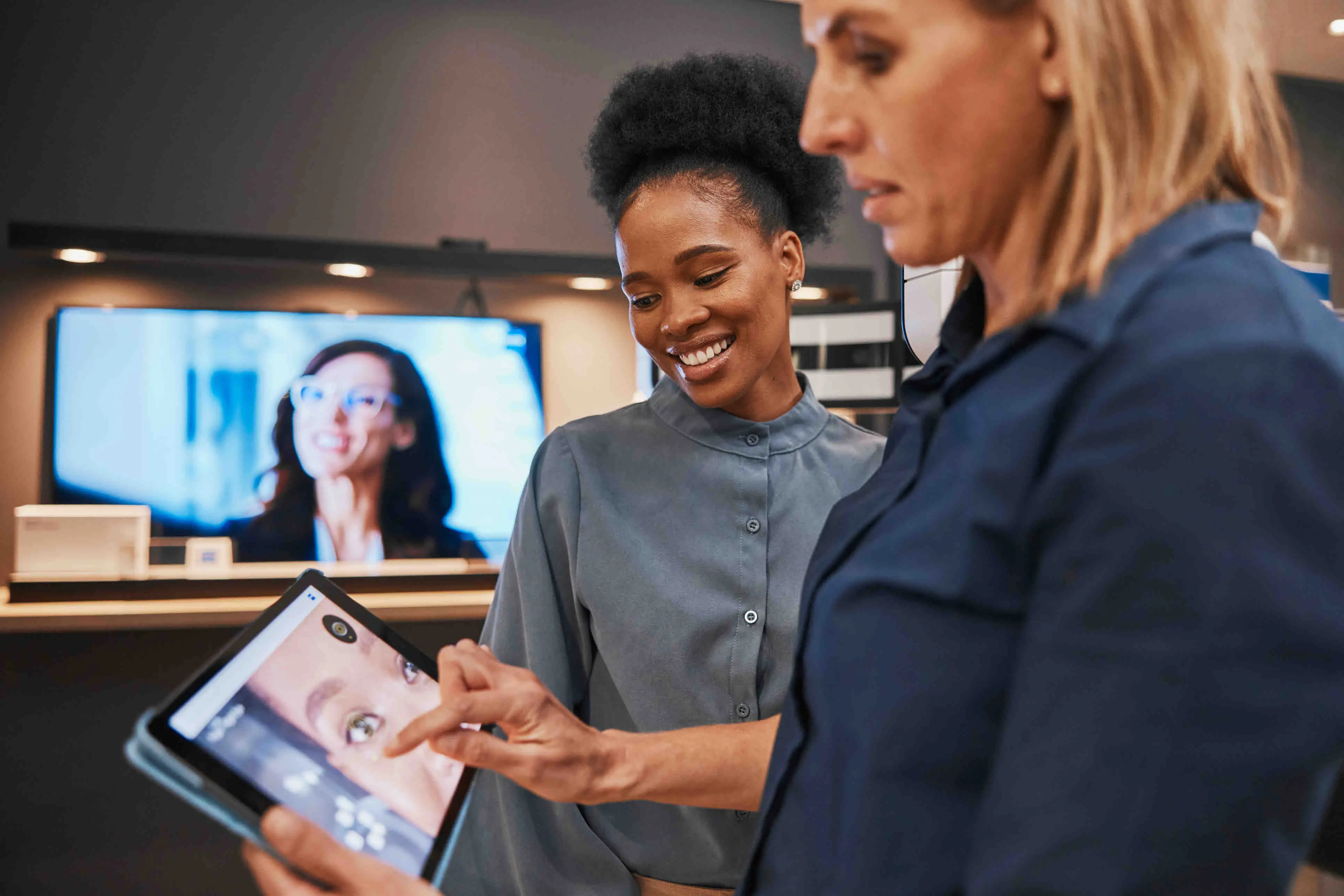 Deux professionnelles discutent en consultant une tablette affichant l’image d’un visage, dans un environnement moderne avec un écran en arrière-plan.