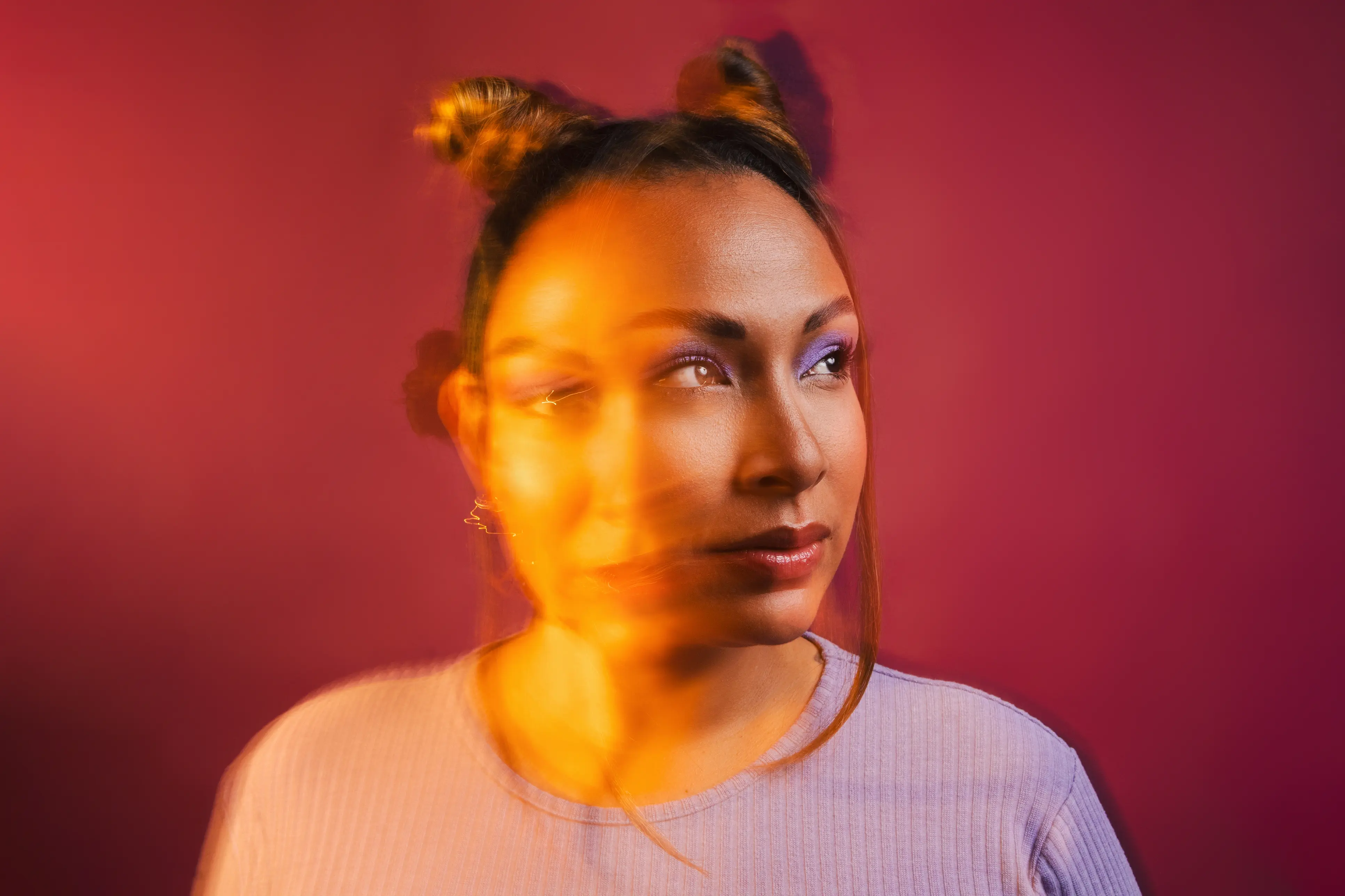 Portrait d’une femme sur un fond rose foncé, regardant sur le côté avec du maquillage violet aux yeux et un effet de lumière orange en mouvement sur son visage.
