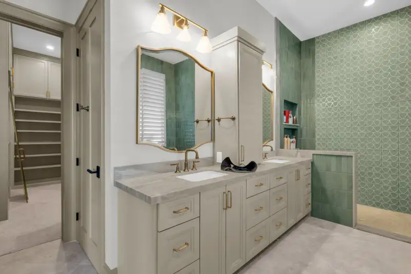 Double vanity with brass faucets, a green tile shower, and a closet at the left.
