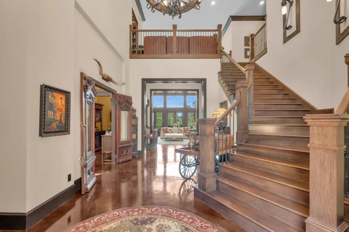Two-story entry hall with a wood staircase, a balcony rail above, polished reddish-brown floors, and a sunlit room beyond.