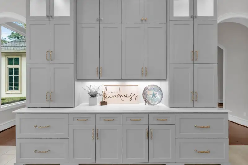 Gray built-in cabinetry with gold pulls, a white countertop, and a lit niche with a decorative sign.