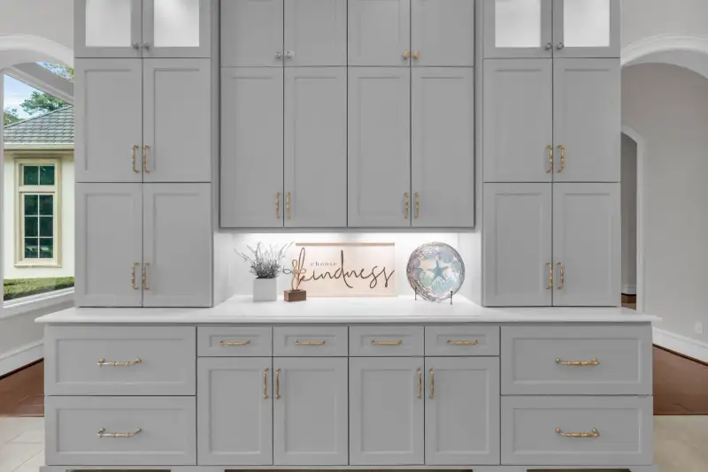 Gray built-in cabinetry with gold pulls, a white countertop, and a lit niche with a decorative sign.