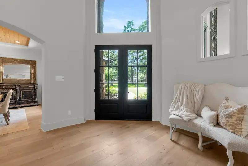 Entry with black French doors opening to a lawn, a bench, and light wide-plank hardwood floors.