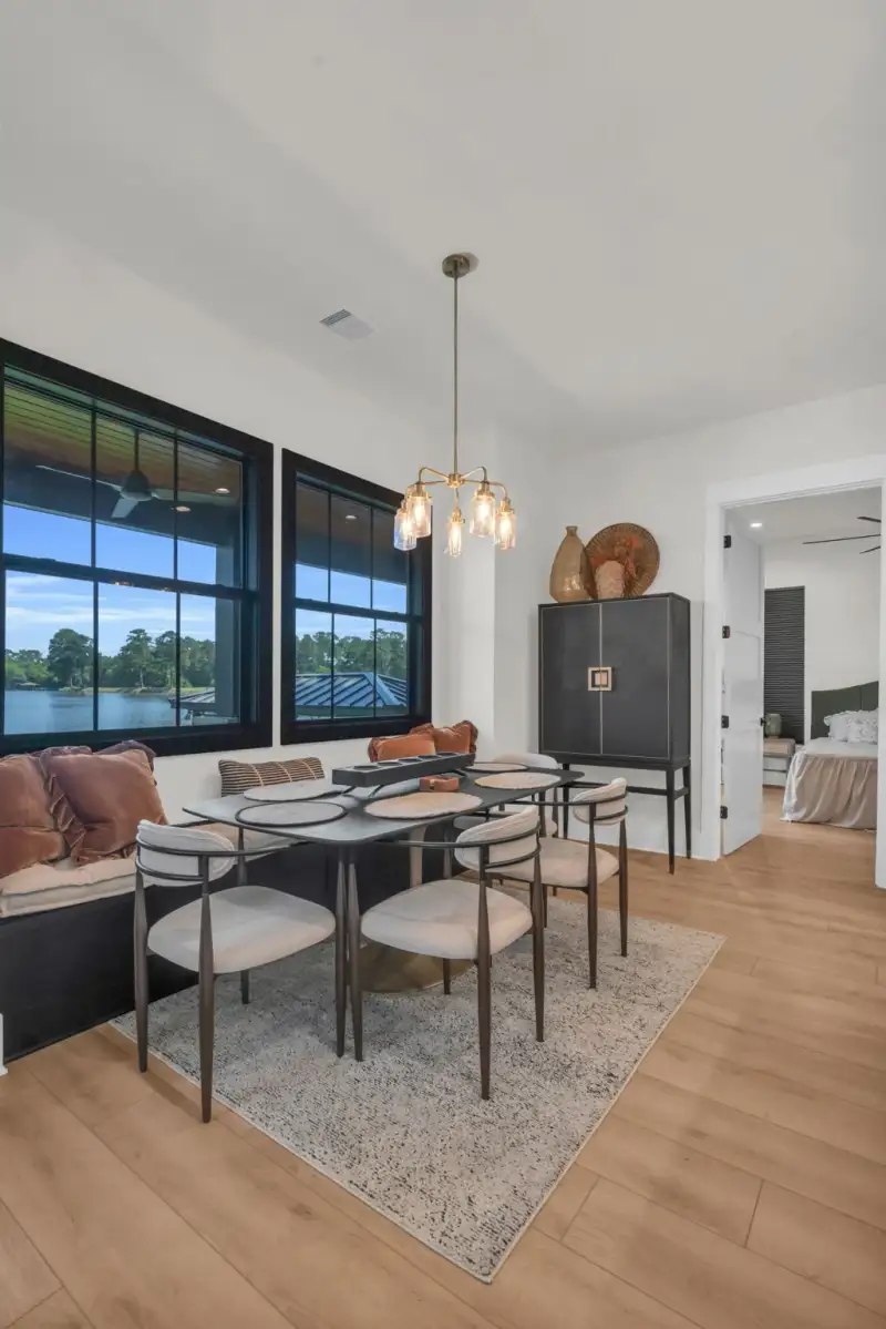 Dining nook with a dark table, upholstered chairs, and black-framed windows overlooking water.