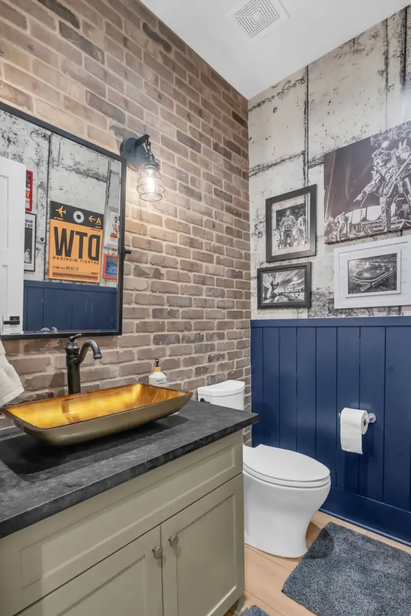 Powder room with exposed brick, a gold vessel sink, a dark countertop, and navy paneling.