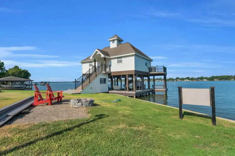 Boathouse on pilings beside the water with red Adirondack chairs and a stone fire pit on the lawn.