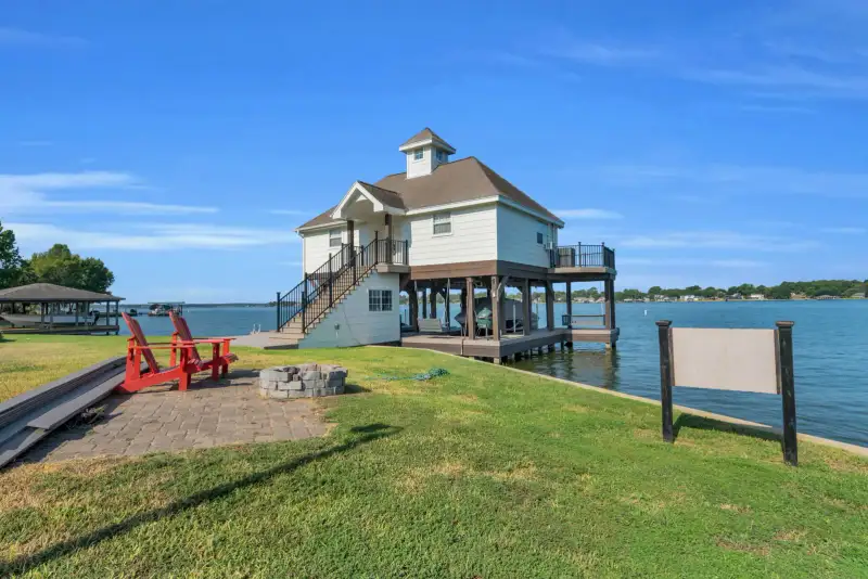 Boathouse on pilings beside the water with red Adirondack chairs and a stone fire pit on the lawn.