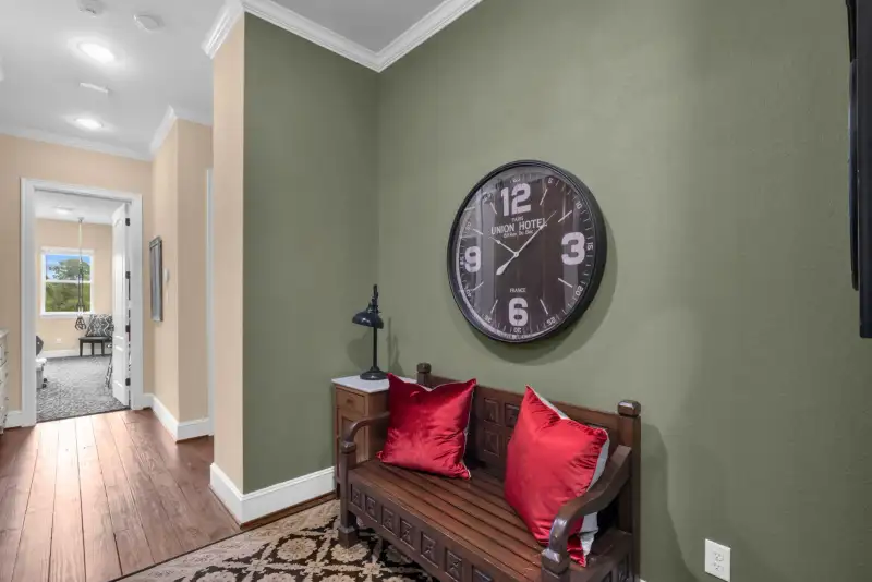 Hallway nook with a green accent wall, a round wall clock, a carved wood bench, and two red throw pillows.