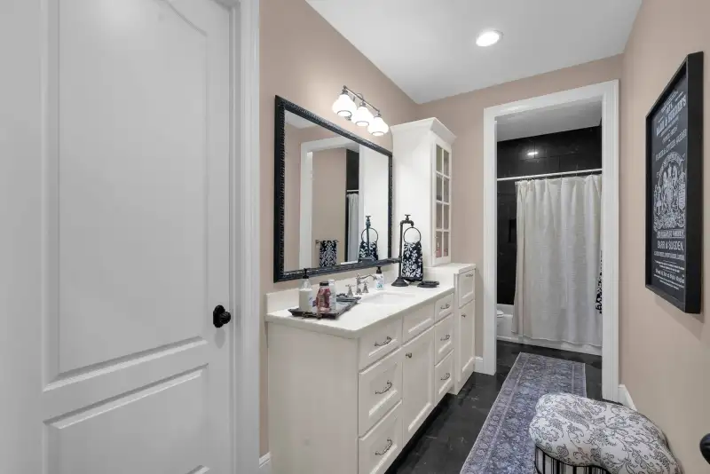 Bathroom with a white vanity, a black-framed wall mirror, a three-light fixture, and a bathtub beyond a doorway.