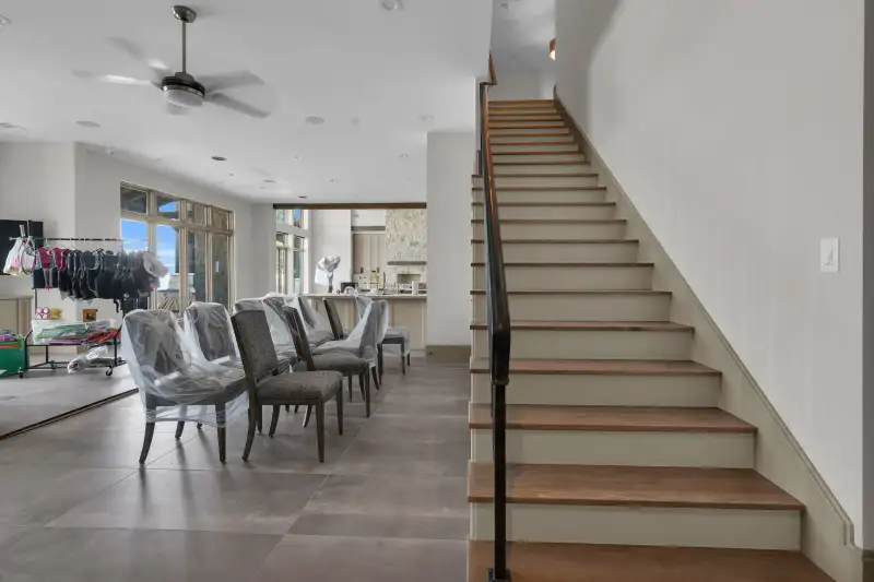 Open room with ceiling fans, dining chairs under plastic, and a wood staircase at the right.