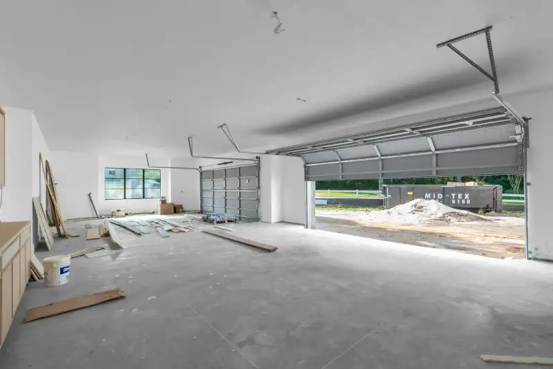 Garage with lumber on the concrete floor and an open overhead door to a yard with a dumpster.