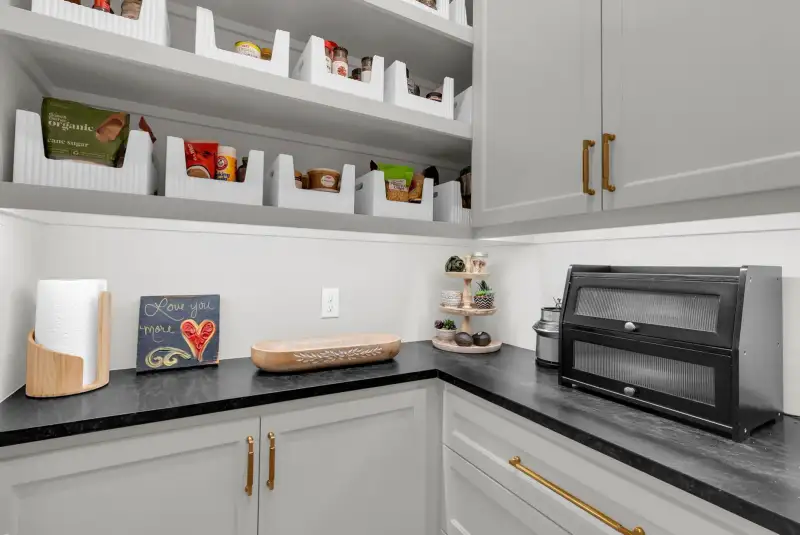 Pantry corner with white bin organizers, a black countertop, and a black bread box with fluted glass front.