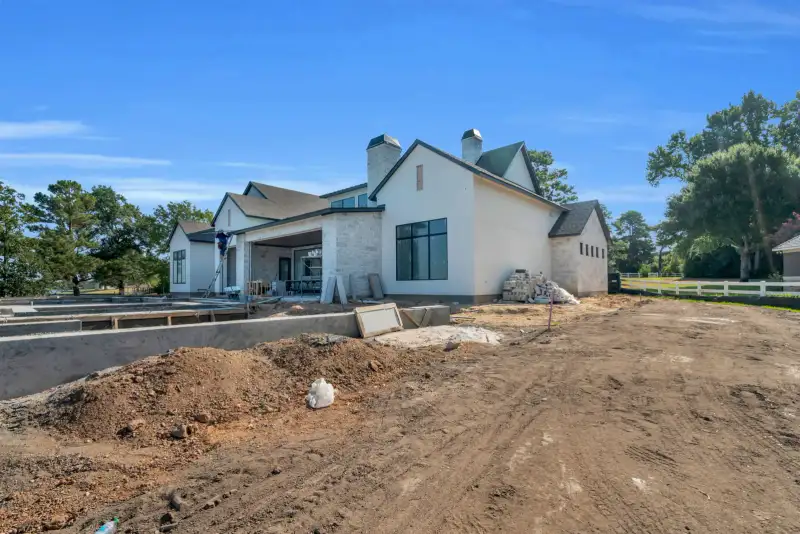 Exterior of a stucco and stone house under construction, with a dirt yard and a white rail fence.