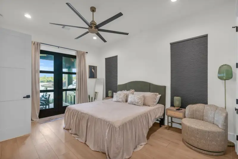 Bedroom with a green upholstered headboard, dark roller shades, a ceiling fan, and glass doors.