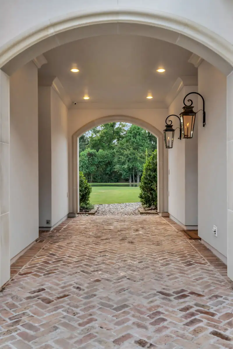 Arched passage with a herringbone brick floor, wall lanterns, and a view through to a lawn.