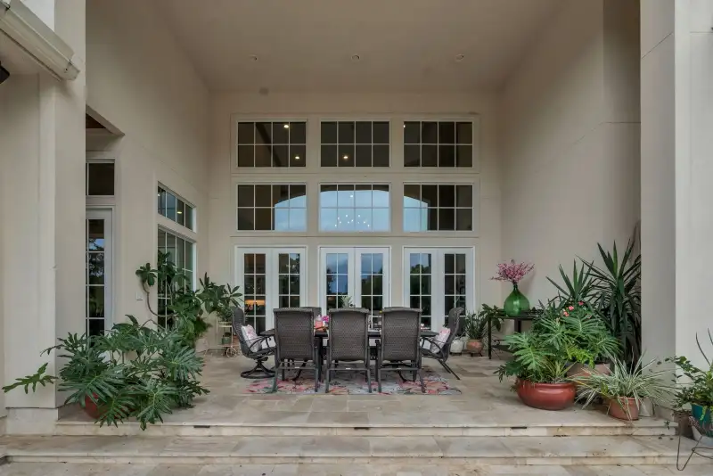 Covered patio dining area with dark wicker chairs, a tall window wall, French doors, and potted plants.