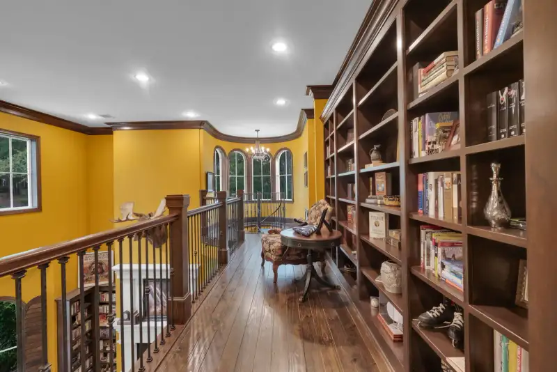 Upper gallery with yellow walls, built-in dark wood bookshelves, a round table, and arched windows at the end.