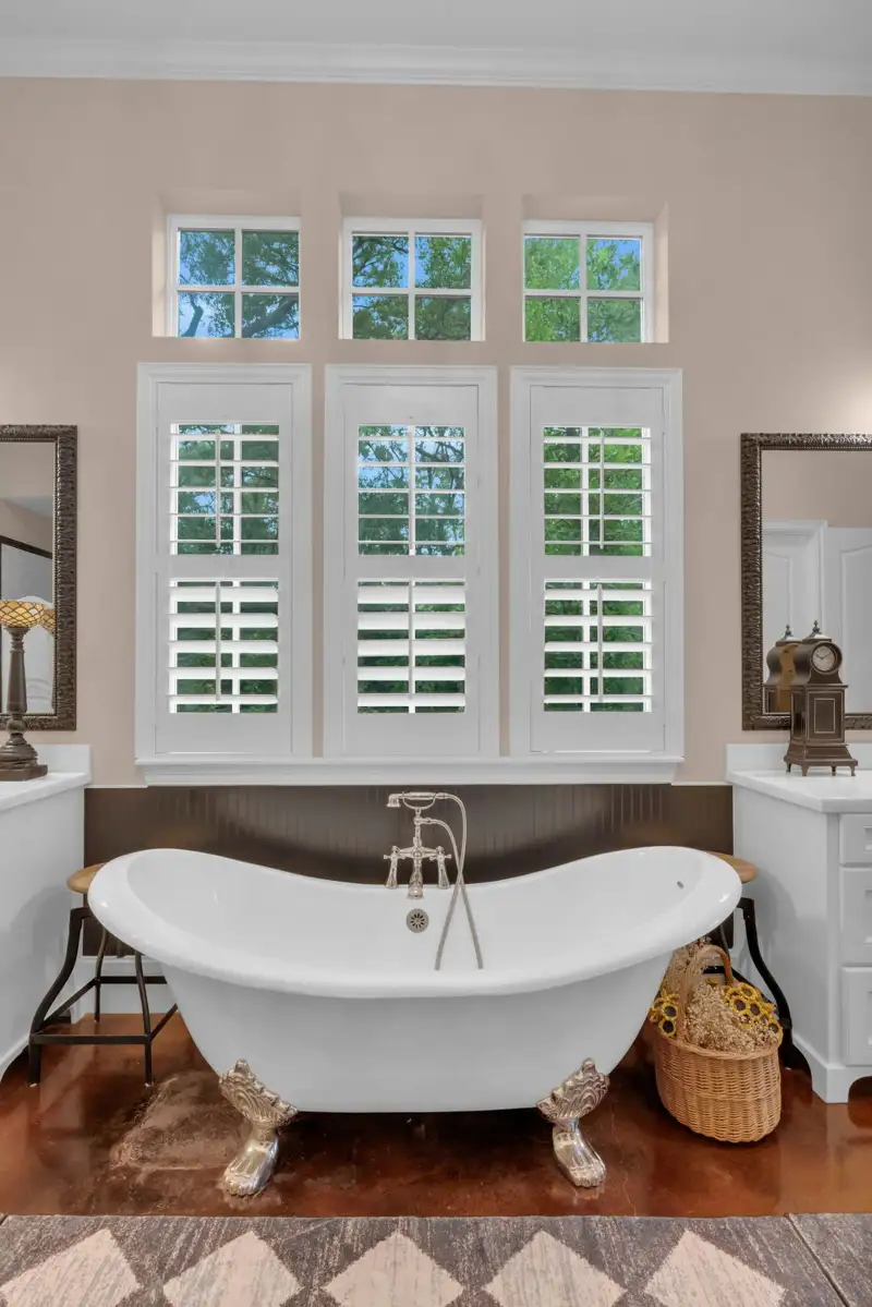 Clawfoot tub beneath three shuttered windows, flanked by white vanity cabinets and a patterned rug.