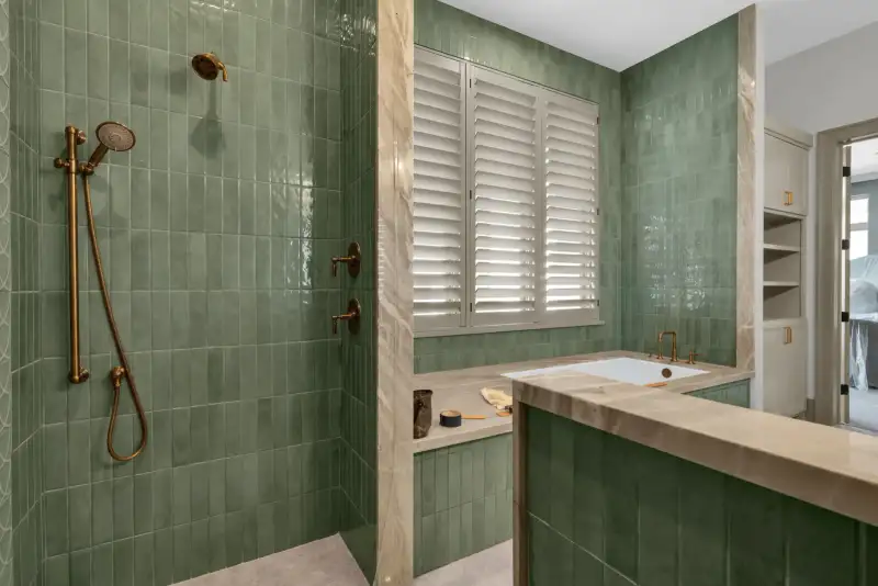 Shower with brass fixtures and green tile, next to a tub with plantation shutters.
