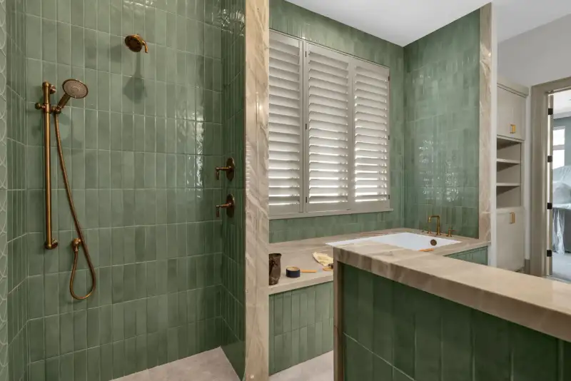 Shower with brass fixtures and green tile, next to a tub with plantation shutters.