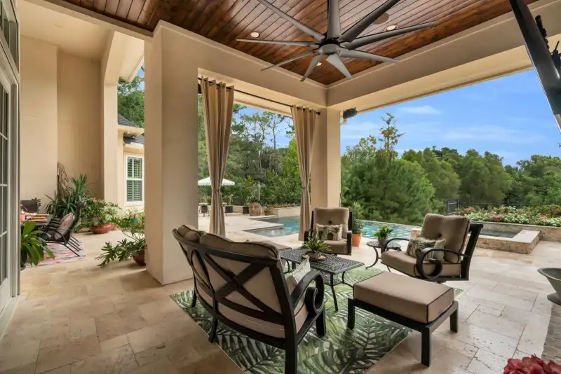 Covered patio with tan cushioned seating, a large wood-planked ceiling fan, and a pool visible beyond curtains.