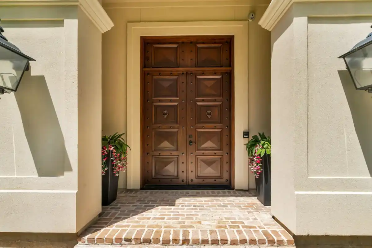 Dark wood double front door with decorative panels, flanked by wall lanterns and potted plants above a brick landing.