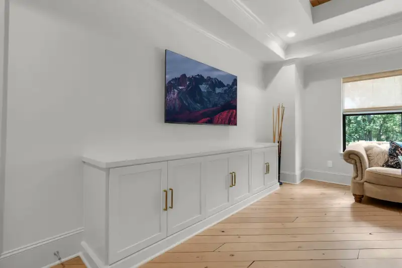 White built-in cabinet under a wall-mounted television, with pool cues standing beside it.