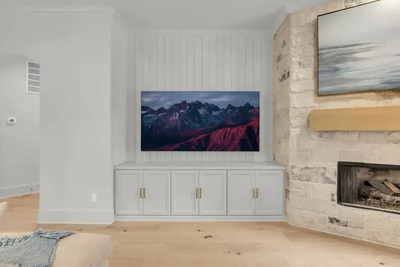 Media wall with a wall-mounted television over built-in white cabinets, beside a stone fireplace.
