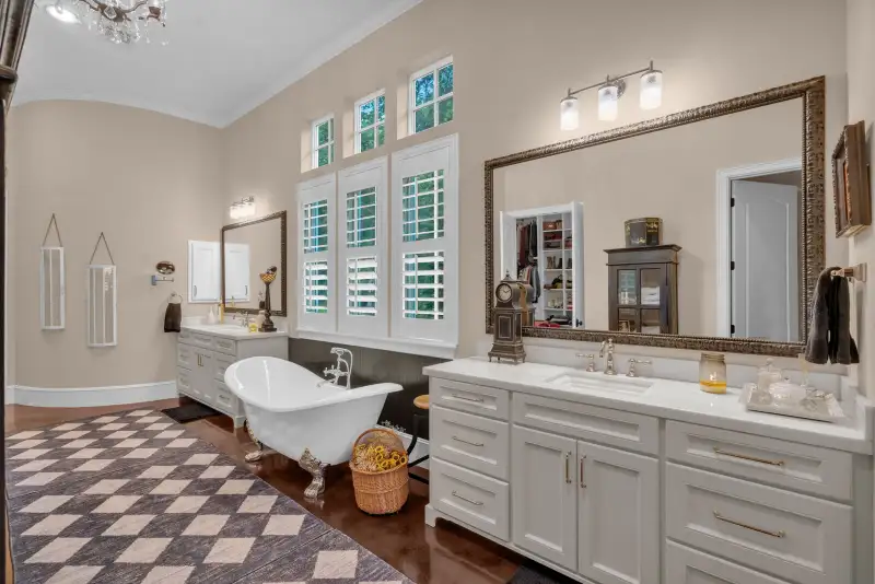 Primary bath with a double vanity, a clawfoot tub, a framed mirror, plantation shutters, and a patterned rug.