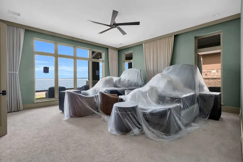 Bedroom with green walls, covered furniture, a ceiling fan, and windows facing the water.