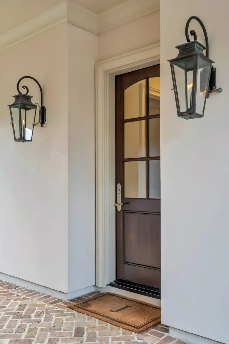 Dark wood entry door with divided glass panels, two wall lanterns, and a welcome mat on brick pavers.