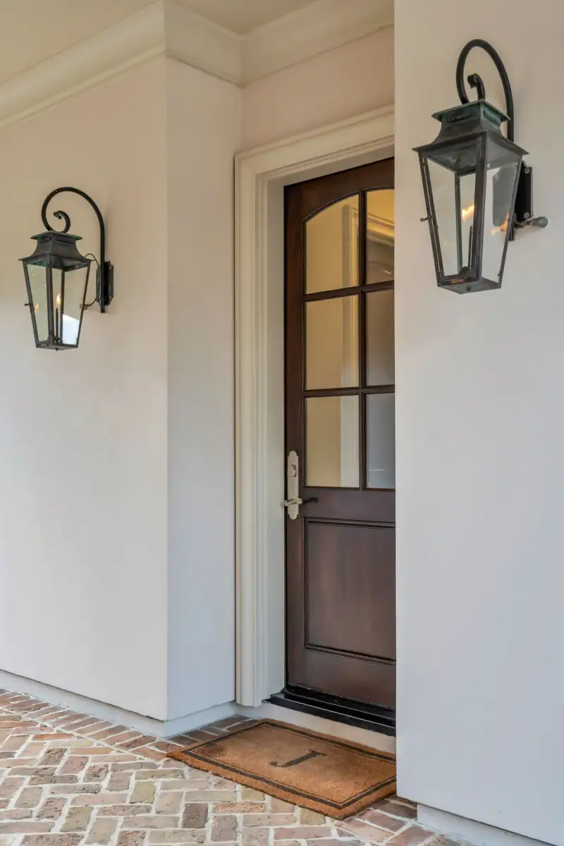 Dark wood entry door with divided glass panels, two wall lanterns, and a welcome mat on brick pavers.