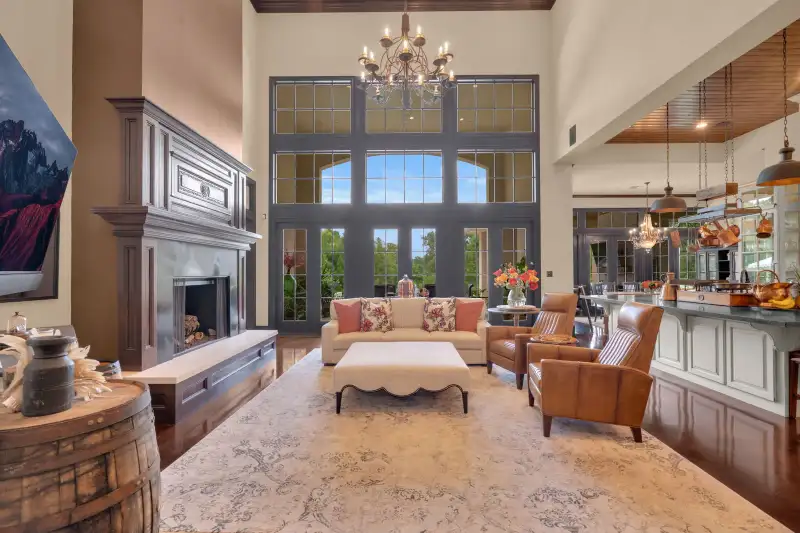 Two-story great room with a gray fireplace, tall windows, a cream area rug, and leather armchairs.