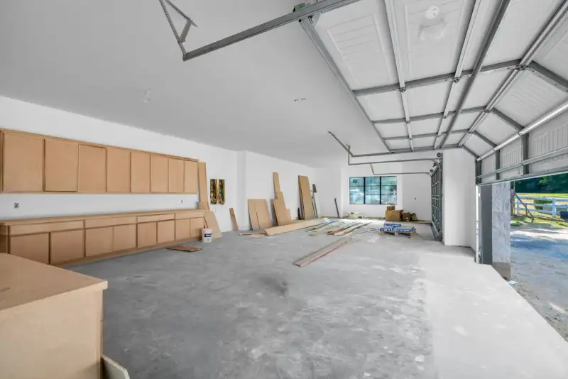 Garage with light wood wall cabinets, bare concrete floor, and an open overhead door at right.