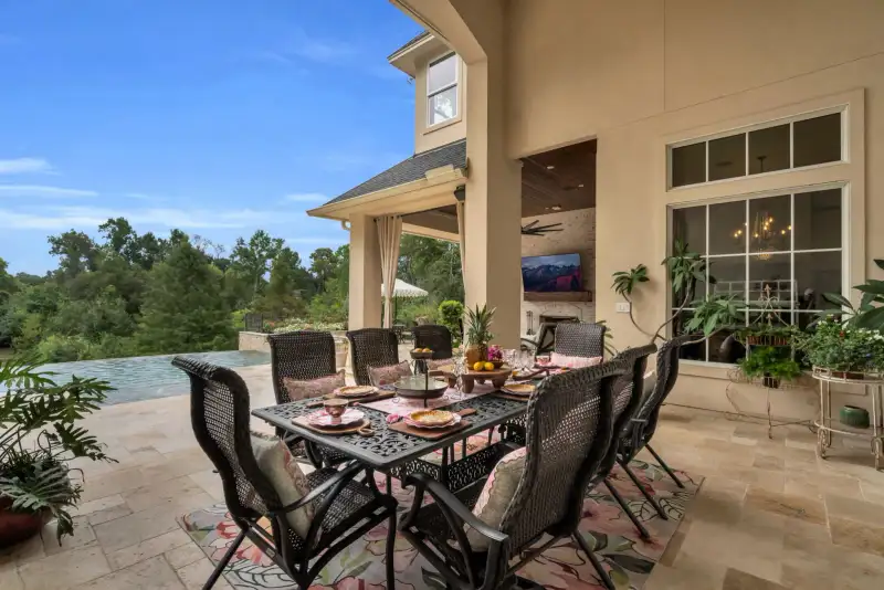 Covered patio with outdoor dining, a stone fireplace with a wall-mounted television, and a pool beyond.