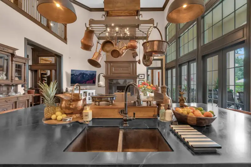 Dark stone kitchen island with a copper sink, a copper pot rack, and a bowl of fruit.