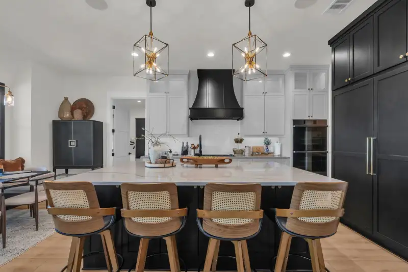 Kitchen island with four woven-back stools under two pendant lights, white cabinets, and a black range hood.
