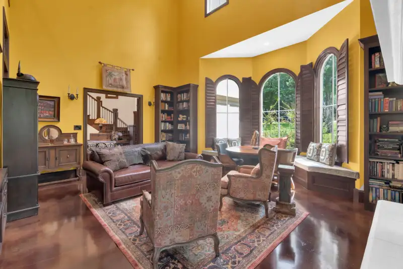 Two-story sitting room with yellow walls, arched windows with wood shutters, a window seat, and a patterned rug.