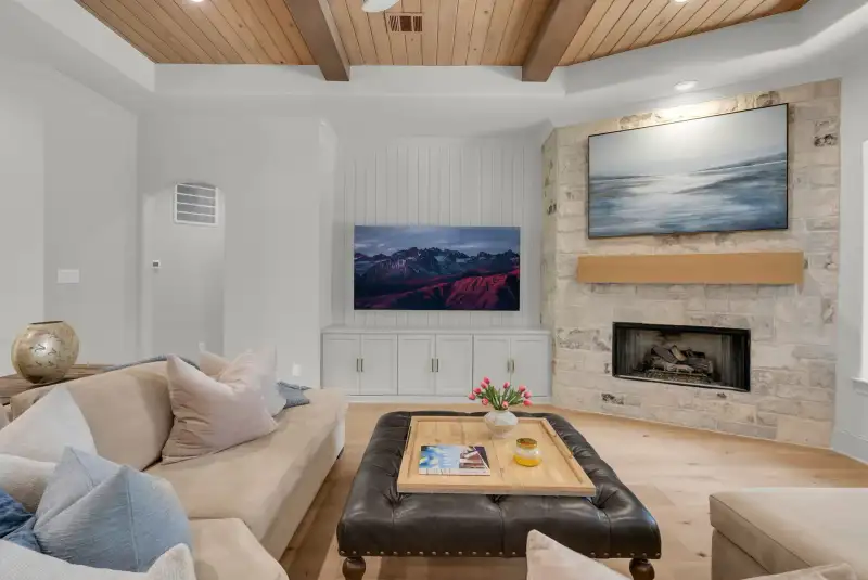 Living room with a stone fireplace, a wall-mounted television above it, and a dark tufted ottoman near a sofa.