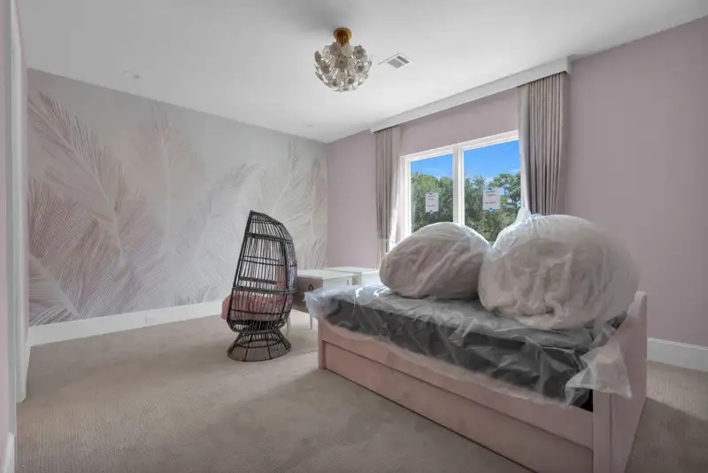 Bedroom with a gray leaf-pattern wall, a wicker accent chair, and a window with trees outside.