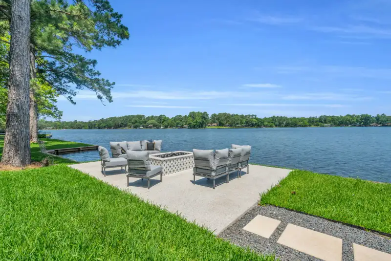 Lakefront patio with gray outdoor seating and a fire pit, with the lake and a shoreline in view.