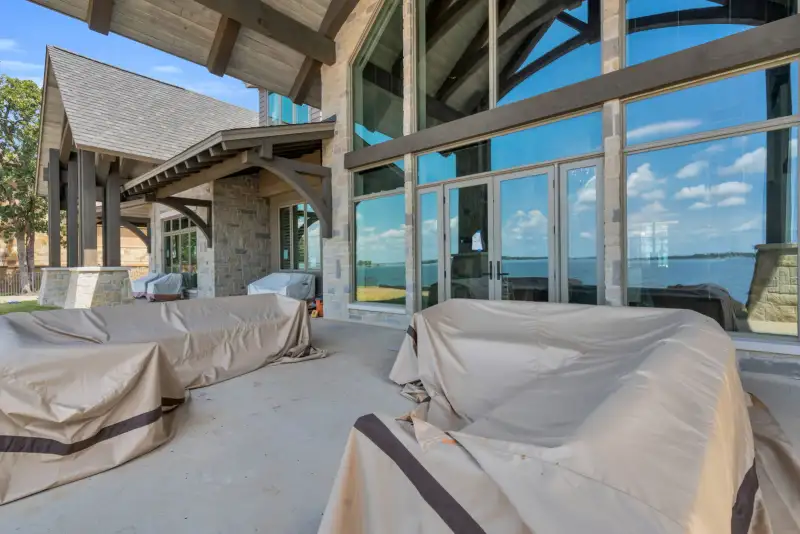 Covered patio with tall glass walls and furniture under covers, with the lake visible through the doors.