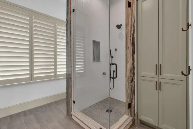 Glass-enclosed shower with plantation shutters on the adjacent window and a tile floor.