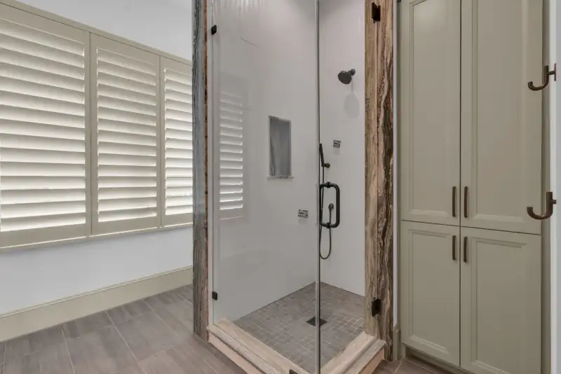Glass-enclosed shower with plantation shutters on the adjacent window and a tile floor.