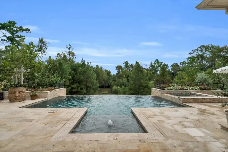 Infinity-edge pool on travertine pavers with a stone planter shaped like a face and trees beyond the fence.
