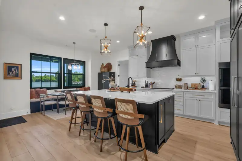 Kitchen and dining nook with a dark island, four woven-back stools, white cabinets, and a black range hood.