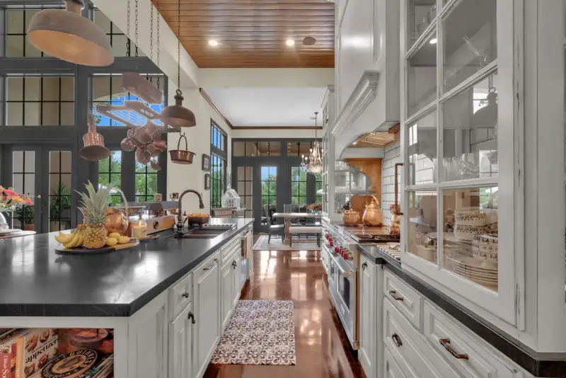 Kitchen island with a dark countertop, a pineapple and fruit display, a copper pot rack, and white glass-front cabinets.